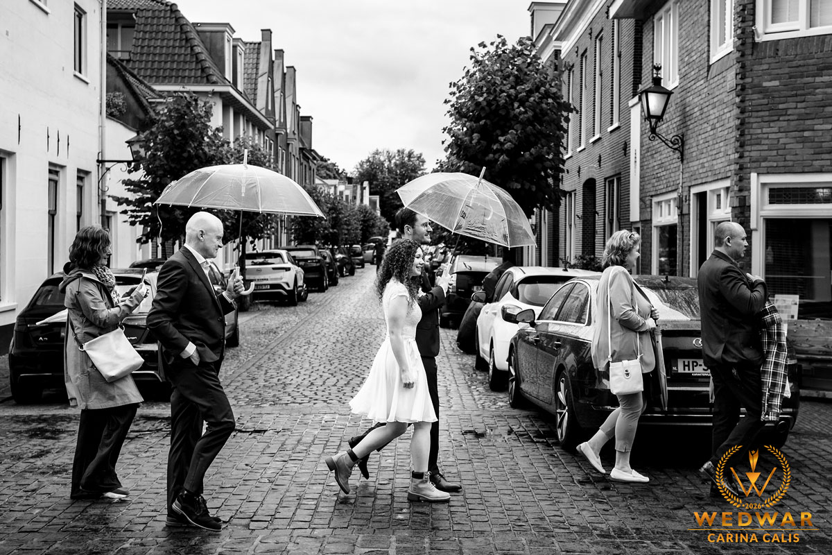 Bride in a white dress walks under a transparent umbrella beside a suited man on a cobblestone street with onlookers and parked cars nearby in a black-and-white street scene and rain-slick pavement.