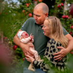Father holding crying baby while mother smiles in an apple orchard setting with fruit trees in the background.