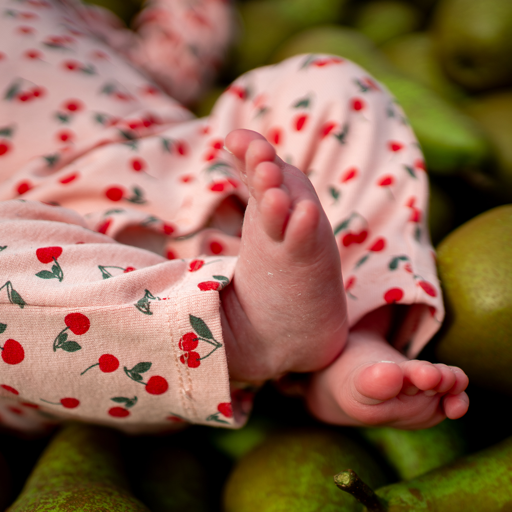 Close-up of a baby's feet resting on a pink cherry-print onesie among green pears.
