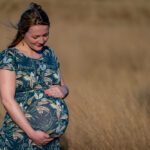 Pregnant woman in a teal leafy-patterned dress stands in a sunlit field, cradling her baby bump with both hands.