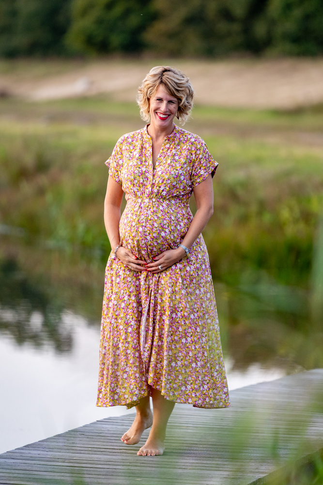 Pregnant woman in a pink floral dress stands barefoot on a wooden dock beside a calm lake, smiling sweetly.