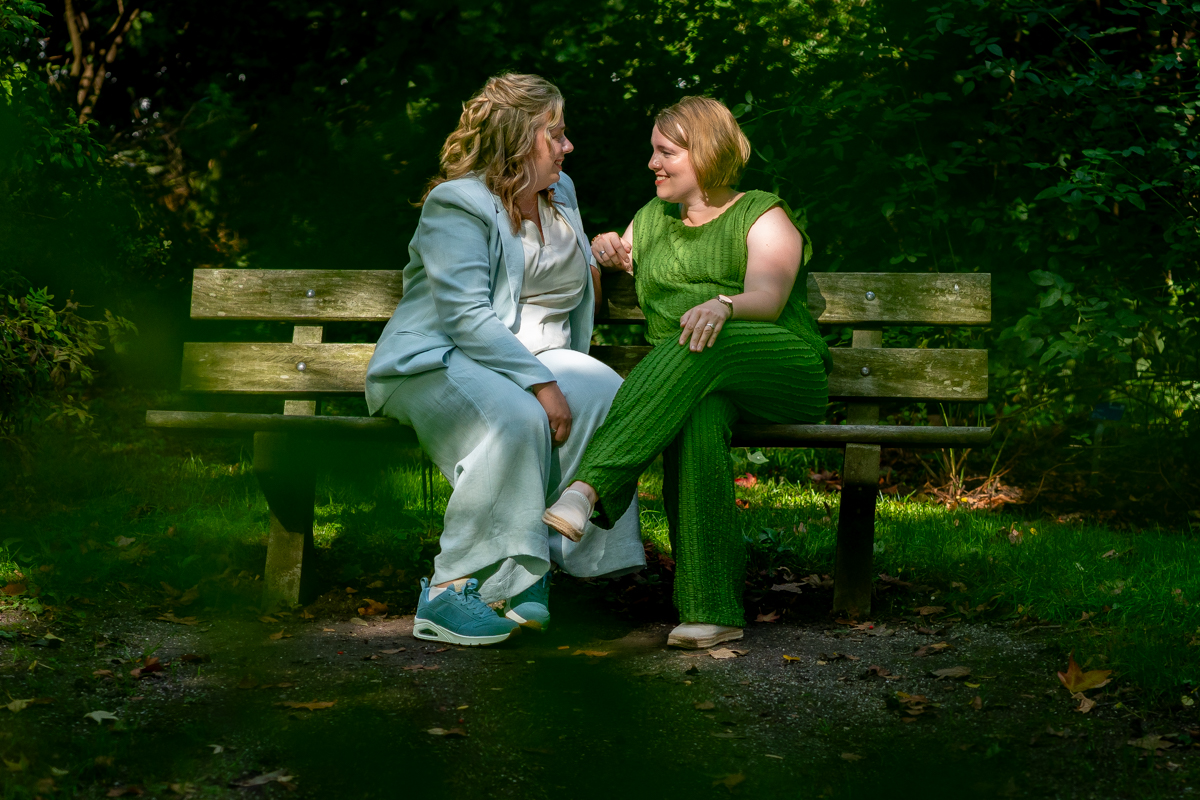 Two women sit on a wooden park bench, smiling and talking amid greenery.