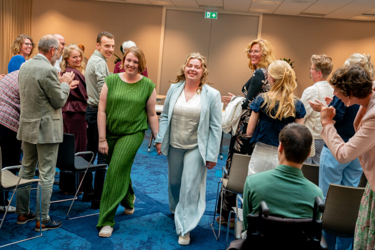 Two women walk down an aisle between seated attendees as everyone smiles and claps at an indoor event.