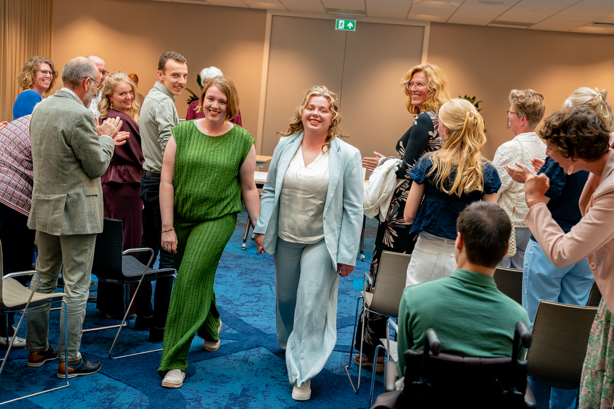 Two women walk down an aisle between seated attendees as everyone smiles and claps at an indoor event.