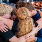 Four people in a close group hug, arms around each other in a supportive embrace in a seating area.